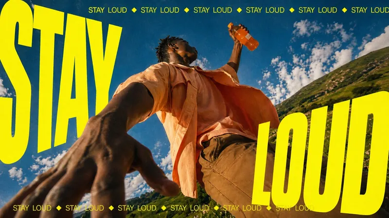 Kinetic type poster: a low-angle shot of a man raising a drink bottle and reaching toward the camera, framed by huge slanted yellow letters STAY LOUD against blue sky and hills (landscape)